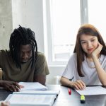 Multiethnic group of classmates sitting at table with notebook and textbook while studying together in university